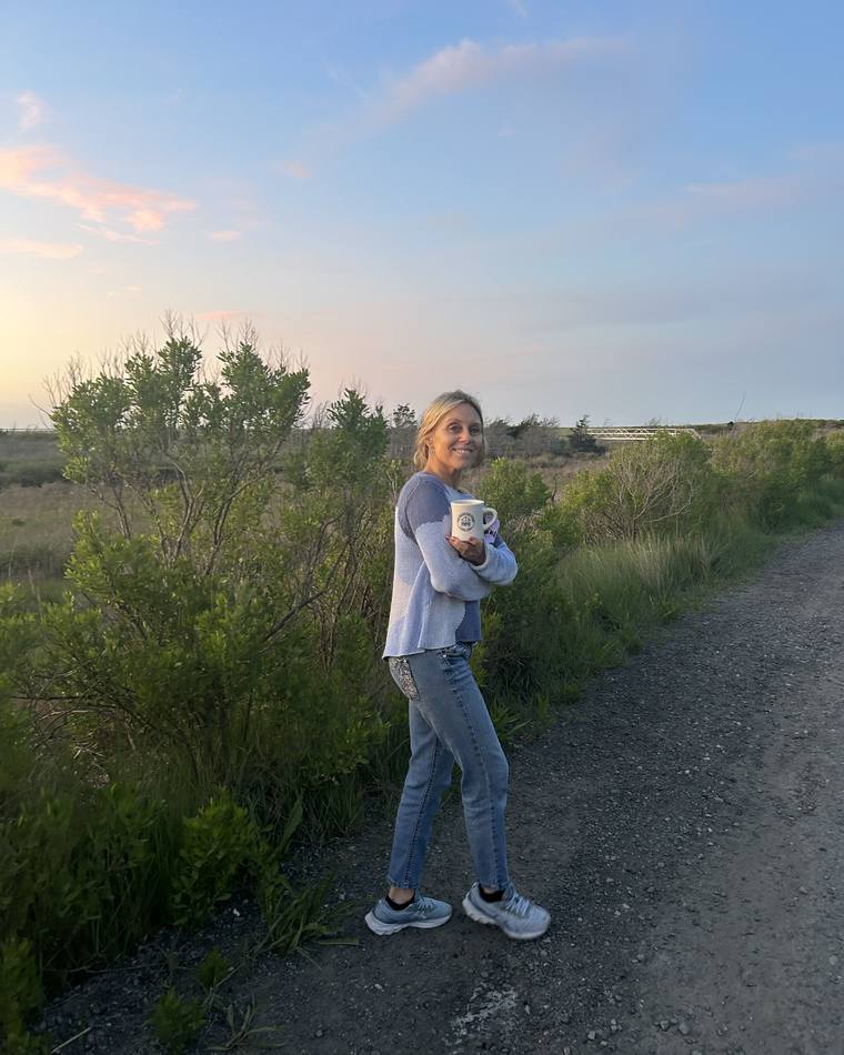 Jill Sylvester outdoors at sunset, holding a mug on a gravel path beside a marsh